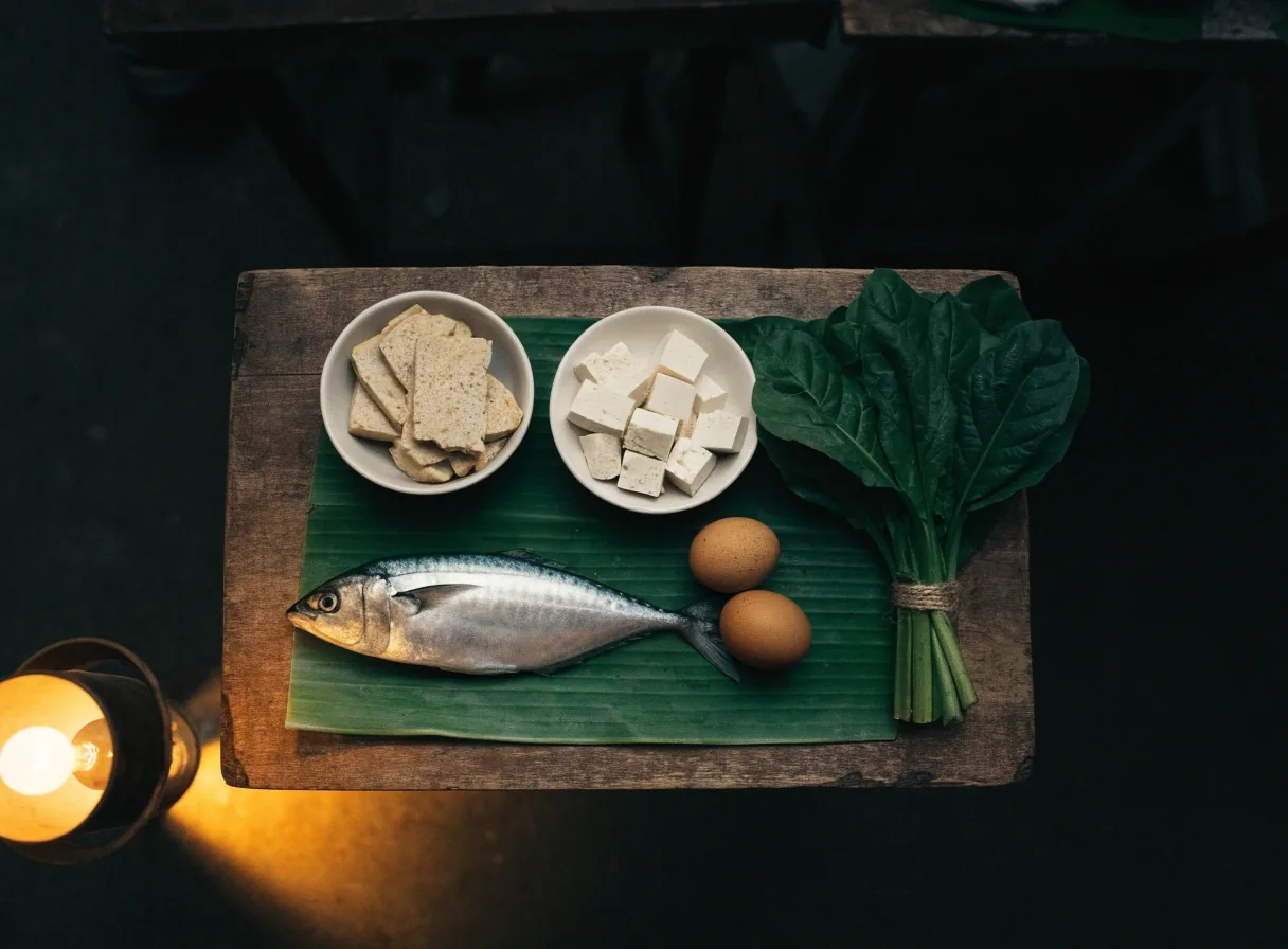 Fresh mackerel, tempeh, tofu, eggs and greens laid out on a market stall table at dusk